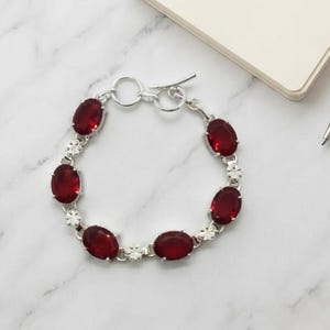 May include: A silver-toned bracelet featuring six oval-shaped, deep red faceted stones linked together. The bracelet includes a circular toggle clasp mechanism at one end and small decorative silver floral accents between each red stone.