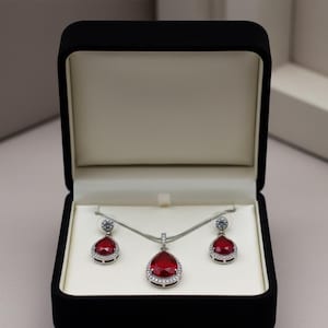 May include: A jewelry set featuring a necklace and earrings with teardrop-shaped, red gemstones. The gemstones are surrounded by a halo of small, clear stones. The set is displayed in an open, black velvet box with a cream-colored interior.