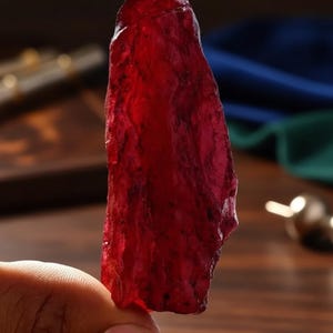 Może przedstawiać: Surowy, półprzezroczysty, głęboko czerwony kamień szlachetny o szorstkim, nieregularnym kształcie. Kamień trzymany jest w dłoni, z rozmytym tłem innych przedmiotów i tkanin. Kamień szlachetny ma bogaty, nasycony czerwony kolor.