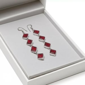 Puede incluir: Pendientes colgantes de plata con cinco gemas rojas cuadradas en cada pendiente, presentados en una caja de joyería blanca con interior gris. Los pendientes están en ángulo.