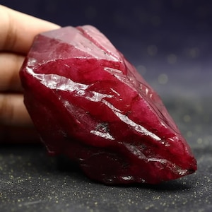 Puede incluir: Una gema de rubí en bruto, sin cortar, de un tono rojo intenso. La superficie de la piedra tiene un aspecto facetado, que refleja la luz. El rubí se sostiene en una mano, mostrando su tamaño y color. Esta gema natural es un ejemplo llamativo de belleza mineral en bruto.