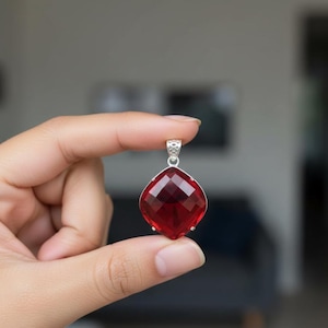 Puede incluir: Un colgante de piedra preciosa roja oscura, tallado en forma cuadrada, con una fianza de plata. El colgante se sostiene entre los dedos, mostrando su talla brillante y su rico color. Una joya llamativa.
