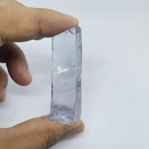 May include: A translucent, rectangular crystal with a light purple hue. The raw, natural edges of the stone are visible, and it is held between two fingers against a white background. The crystal's surface reflects light.