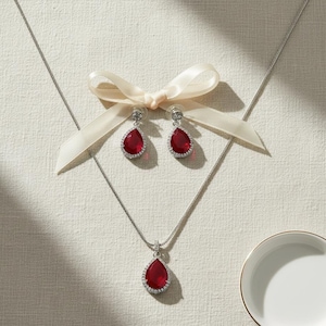 May include: A silver necklace and matching earrings set with teardrop-shaped, ruby-red gemstones. The gemstones are surrounded by a halo of small, clear crystals. The earrings are displayed with a cream-colored ribbon.