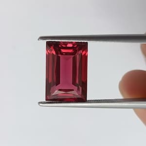 Puede incluir: Una gema rectangular facetada sujeta con pinzas. La gema es de un color rojo intenso con una superficie pulida. El corte es de esmeralda, con facetas paralelas. La gema se sostiene sobre un fondo blanco.