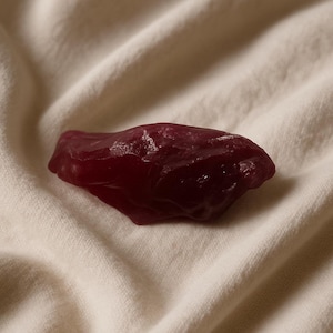 Puede incluir: Una piedra de color rojo oscuro, de forma irregular y con una superficie ligeramente brillante. La piedra descansa sobre una tela crema texturizada, creando un contraste de color y textura. La forma de la piedra es orgánica y natural.