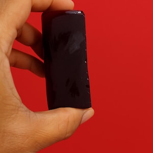 May include: A dark purple, rectangular object with a glossy finish is held against a red background. The object appears to be a solid, possibly a bar or block, with a slightly irregular edge. The lighting creates highlights and shadows on its surface.