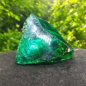 Puede incluir: Una gran pieza de vidrio verde vibrante de forma irregular, posiblemente un objeto decorativo. El vidrio es translúcido, permitiendo el paso de la luz y creando un efecto brillante. El objeto descansa sobre una superficie oscura y texturizada. Es probable que sea un artículo decorativo.