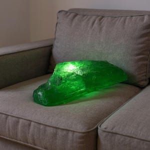 以下が含まれることがあります： 大きな、不規則な形の、輝く緑色の装飾品で、宝石に似ています。灰色の布張りのソファの上に置かれています。内部の光源が、テクスチャ加工された表面を照らしています。