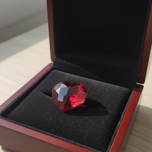 Könnte beinhalten: Ein facettierter, herzförmiger, roter Edelstein liegt in einer offenen Holzschatulle mit schwarzem Samtfutter. Die Schatulle hat eine dunkelrote Oberfläche, die den Glanz des Edelsteins hervorhebt.