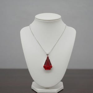 Könnte beinhalten: Eine silberne Halskette mit einem großen, facettierten, roten Anhänger in Tropfenform. Der Anhänger wird auf einer weißen Schmuckbüste vor neutralem Hintergrund präsentiert. Die Halskette ist ein Schmuckstück.
