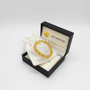 Op de afbeelding: Een goudgeel kralenarmbandje ligt in een zwarte doos, samen met een wit zakje met trekkoord. De doos bevat een laboratoriumgecertificeerde kaart. De armband is gemaakt van ronde, doorschijnende kralen.