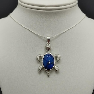 Può includere: Una collana argentata con un ciondolo a forma di tartaruga. Il ciondolo ha una pietra blu a forma ovale incastonata in una cornice a forma di tartaruga. La testa, le pinne e la coda della tartaruga sono dettagliate.