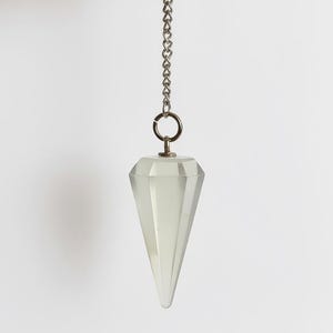 Puede incluir: Un péndulo de cristal claro y facetado cuelga de una cadena plateada. El péndulo es de un color claro y translúcido y se estrecha hasta un punto. La cadena está unida a un pequeño anillo en la parte superior.