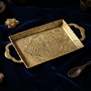 May include: A rectangular gold-colored metal tray featuring intricate embossed floral and geometric patterns across its surface. The tray includes two decorative handles on opposite ends, each with a scalloped edge design and textured detailing.