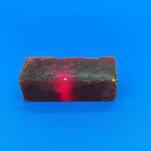 May include: A rectangular, dark red object with a small, bright red light emanating from its center. The object appears to be sitting on a solid blue surface. The texture of the object is slightly rough.