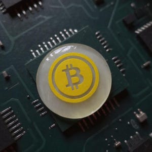 Handmade clear and yellow 2-inch physical Bitcoin coin paperweight resting on a wooden trading desk beside a computer monitor.