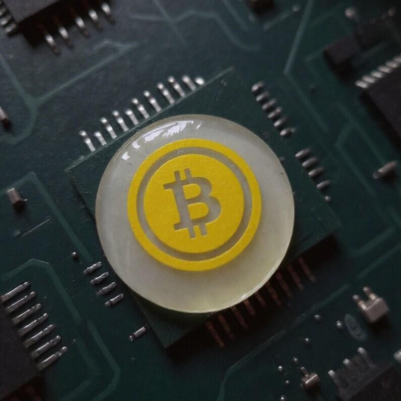 Handmade clear and yellow 2-inch physical Bitcoin coin paperweight resting on a wooden trading desk beside a computer monitor.