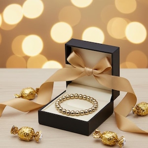 以下が含まれることがあります： 金色のビーズブレスレットが、金色のリボンが付いた黒いギフトボックスに入っています。 金色のリボンと包まれたキャンディーが箱の周りに配置され、お祝いのディスプレイを作り出しています。 背景には、柔らかくぼやけた金色の光があります。