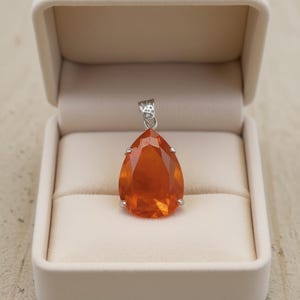 May include: A teardrop-shaped orange gemstone pendant with a silver bail, set in a white jewellery box. The gemstone has a faceted surface, catching the light. The box is open, revealing a soft, cream-coloured interior.
