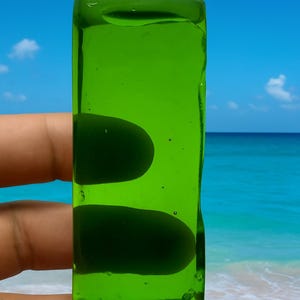 Op de afbeelding: Een rechthoekig, doorschijnend, heldergroen glazen object. Het object wordt omhoog gehouden tegen een achtergrond van een blauwe lucht, oceaan en zandstrand. Het glas heeft een glad oppervlak met afgeronde randen.