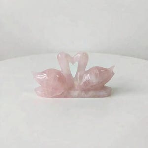Rose quartz love birds figurine displayed on a clean surface next to green foliage, emphasizing its role in relationship healing and marriage harmony.