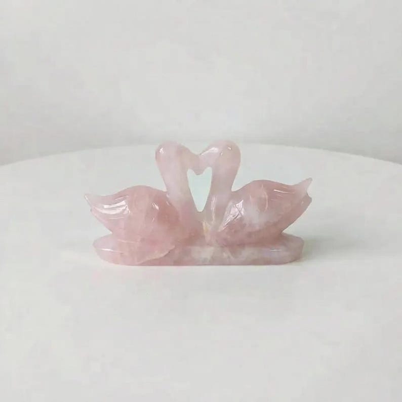 Rose quartz love birds figurine displayed on a clean surface next to green foliage, emphasizing its role in relationship healing and marriage harmony.