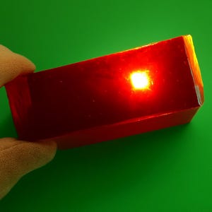 May include: A rectangular, translucent, red gemstone with a bright, glowing center. The gemstone is held against a green background, showcasing its polished surfaces and internal light refraction. The edges of the stone are slightly illuminated.