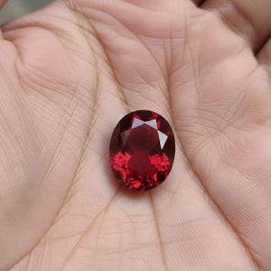 Puede incluir: Una gema roja oscura, facetada y de forma ovalada. La gema se sostiene en la palma de una mano abierta, mostrando su talla brillante y su rico color. Las facetas de la gema reflejan la luz, creando un efecto brillante.