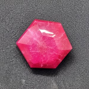 May include: A faceted, hexagonal gemstone in a vibrant shade of pink. The gem has multiple facets that catch the light, creating a sparkling effect. The stone's surface displays a natural, textured pattern.