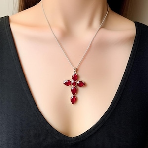 Pode incluir: Um colar de prata com um pingente de cruz. A cruz é adornada com várias pedras preciosas vermelhas ovais e em forma de gota. O colar é usado sobre uma blusa preta.