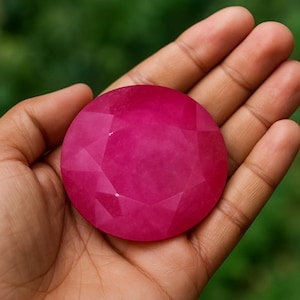 Puede incluir: Una gema grande, facetada y redonda de color rubí. La gema se sostiene en una mano, mostrando su superficie pulida y sus intrincados cortes. El tono rojo intenso de la piedra destaca sobre el tono de la piel, con un fondo verde borroso.