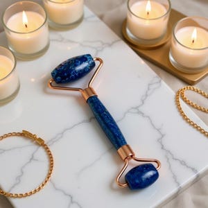 Puede incluir: Un rodillo facial azul y cobrizo con un mango veteado y dos rodillos de piedra azul. El rodillo está sobre una superficie de mármol blanco, con velas encendidas en portavelas de cristal y una pulsera y collar dorados.