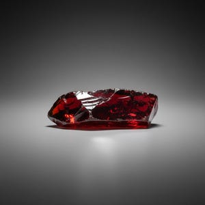 May include: A raw, uncut gemstone with a deep red hue. The rectangular stone has a rough, uneven surface with sharp edges, reflecting light. The gem is set against a gradient gray background, highlighting its natural beauty. This is a gemstone.