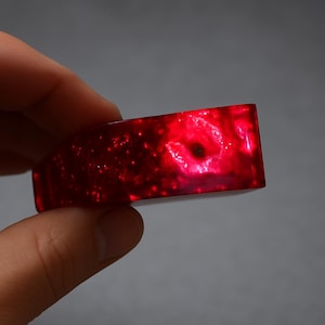 Peut inclure: Un bracelet rectangulaire rouge translucide. Le bracelet présente un motif semblable à une galaxie avec de petites inclusions scintillantes. La lumière traverse le bracelet, créant un effet lumineux. Le bracelet est tenu par une main.