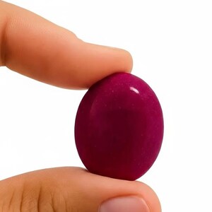 Könnte beinhalten: Ein glattes, ovales Objekt in tiefem Magenta wird zwischen zwei Fingern vor weißem Hintergrund gehalten. Die glänzende, reflektierende Oberfläche deutet darauf hin, dass es aus poliertem Stein oder Glas besteht. Die Form ist schlicht.