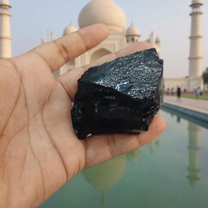 May include: A hand displays a piece of black obsidian, a volcanic glass with a shiny surface. The stone's irregular shape is prominent. The Taj Mahal, a white marble mausoleum, is in the background, reflected in the water.