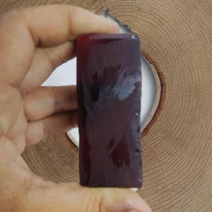 May include: A rectangular, translucent, dark red object is held in a hand. The object has a smooth surface with some internal marbling. The background is a light brown, circular wooden surface with a hole in the center.