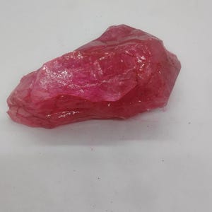 May include: A raw, uncut ruby gemstone with a vibrant, deep red hue. The natural stone has a rough, irregular shape and a slightly translucent appearance, reflecting light across its surface. This gemstone is a beautiful example of natural mineral formations.