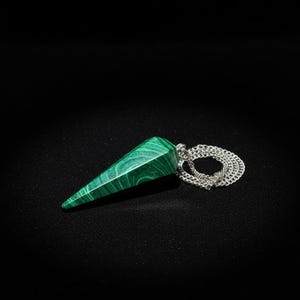 May include: A green malachite pendulum with a silver chain. The pendulum is a pointed, elongated shape with a polished surface. The malachite has a distinctive pattern of swirling green and darker green bands.