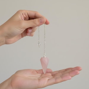 May include: A rose quartz pendulum with a silver chain and a clear crystal bead. The pendulum is held between two hands against a neutral background. The rose quartz is a pale pink colour and is cut into a pointed shape.