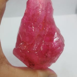 Könnte beinhalten: Ein roher, ungeschliffener Rubin-Edelstein. Der Rubin hat eine tiefe, lebendige, rötlich-rosa Farbe mit einer kristallinen Struktur. Der Edelstein wird in einer Hand vor weißem Hintergrund gehalten.