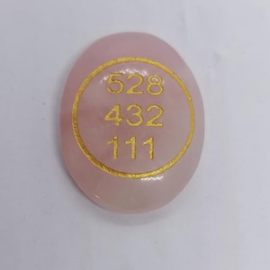 May include: A smooth, oval rose quartz stone. It features the gold numbers 528, 432, and 111 within a gold circle. The stone is a soft pink colour and appears polished.