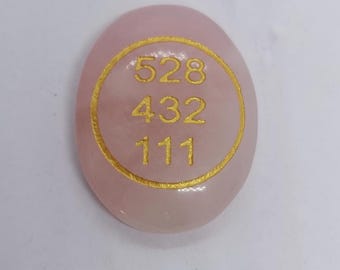 Puede incluir: Una piedra de cuarzo rosa lisa y ovalada con los números dorados 528, 432 y 111 dentro de un círculo dorado. La piedra es de color rosa suave y parece pulida.