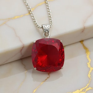 Könnte beinhalten: Eine silberne Halskette mit einem großen, quadratischen, rubinroten Anhänger. Der Anhänger ist in einen silbernen Rahmen gefasst und hängt an einer feinen Silberkette. Der Edelstein hat einen brillanten Glanz und zieht die Blicke auf sich.