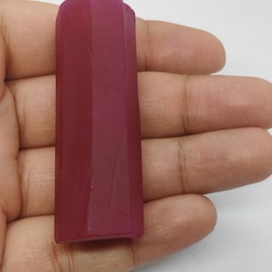 Op de afbeelding: Een gefacetteerde, rechthoekige, diep magenta gekleurde edelsteen. De steen wordt in een hand gehouden tegen een witte achtergrond. De edelsteen heeft een gepolijst, glad oppervlak en een geometrische vorm.