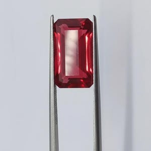 Peut inclure: Une gemme rouge foncé, taillée en rectangle, est maintenue par une pince argentée sur fond blanc. L'intérieur de la gemme est plus clair, également rectangulaire. Il s'agit probablement d'un rubis ou d'un grenat.