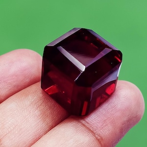 Puede incluir: Una gema facetada de color rojo intenso con forma geométrica. La gema se sostiene sobre un fondo verde, mostrando sus superficies pulidas y su rico color. Las facetas de la gema reflejan la luz, creando un efecto brillante.