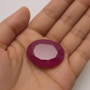 Könnte beinhalten: Ein facettierter, ovalförmiger, tiefroter Edelstein. Der Edelstein wird in einer Hand gehalten, wodurch seine polierte Oberfläche und geometrischen Schnitte hervorgehoben werden. Die Farbe des Steins ist ein satter, gesättigter Farbton, der das Licht über seine vielen Facetten reflektiert.