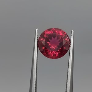 Puede incluir: Una gema redonda, facetada, de color rojo intenso, sujeta con pinzas plateadas. La gema tiene un patrón de estrella en el centro y se encuentra sobre un fondo gris. La piedra preciosa es de un tono rojo vibrante.
